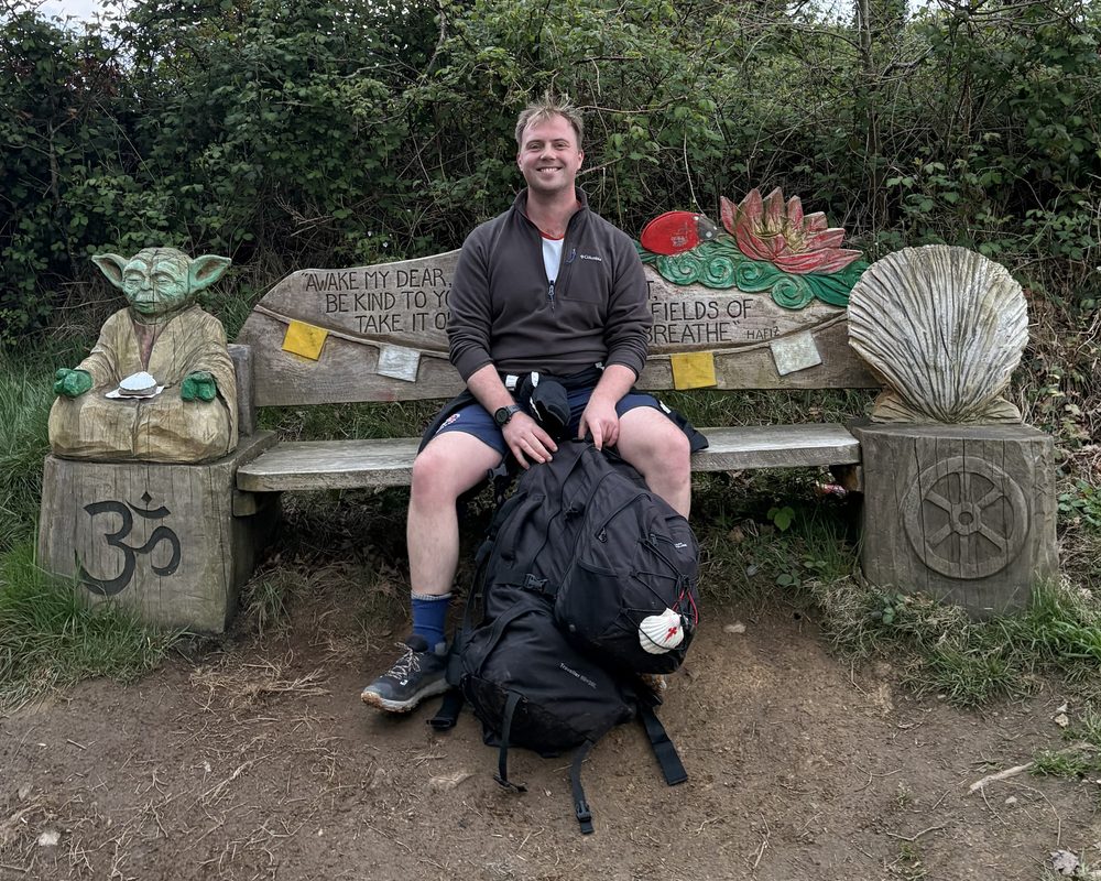 Rhys smiling on a carved wooden bench, between a wooden Yoda and a carved scallop shell